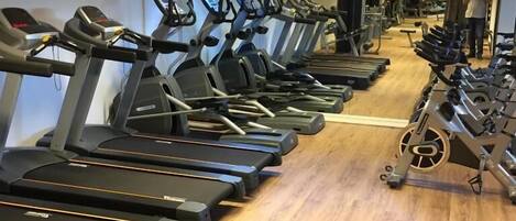 A row of treadmills in a gym with wooden flooring and large windows.