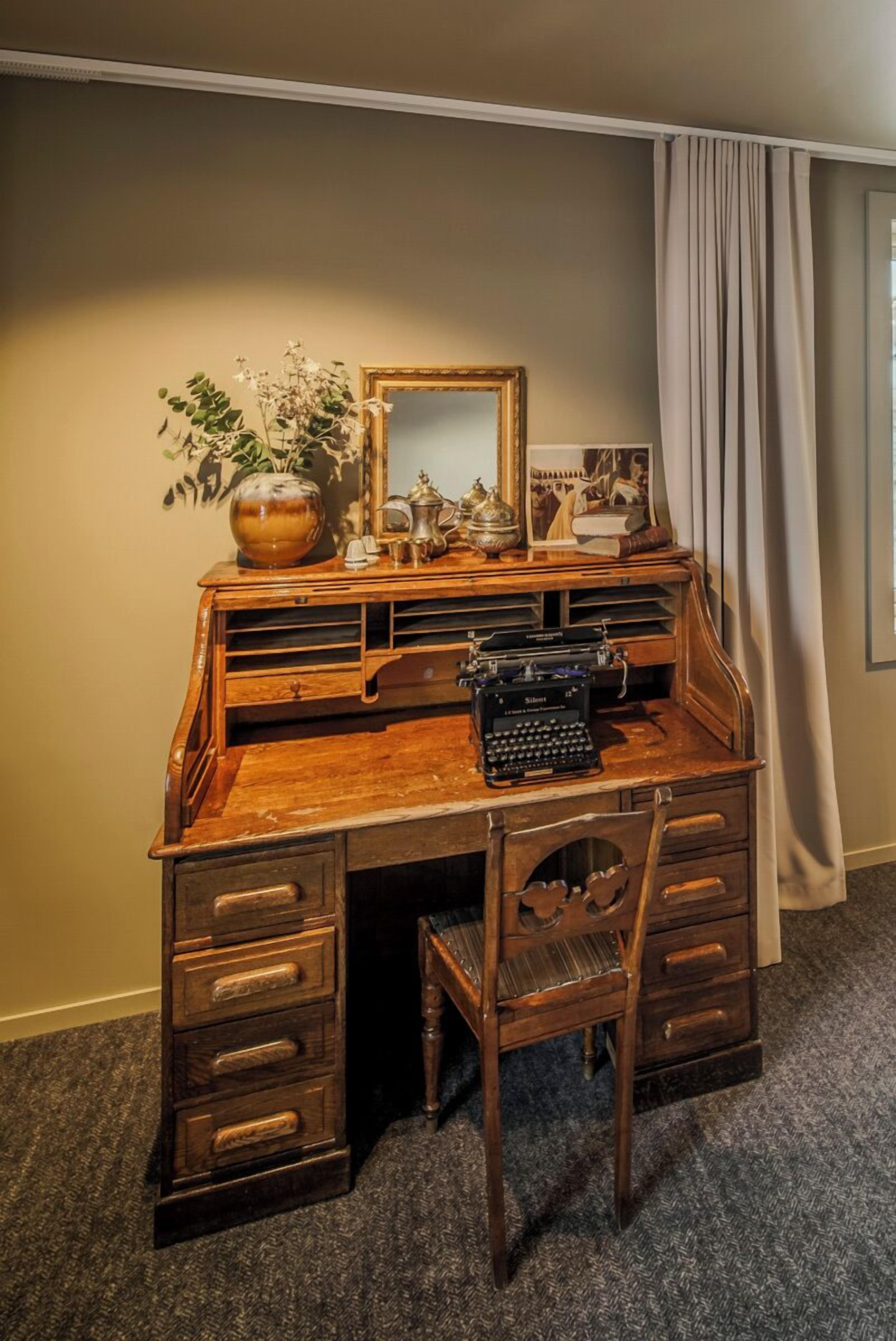 A wooden desk with a vintage typewriter, a vase with dried flowers, a framed picture, and a mirror.