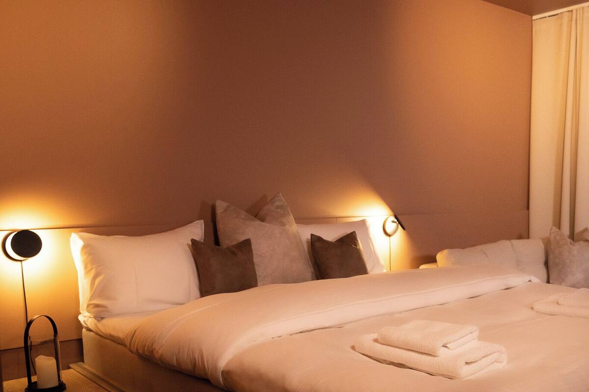 Ein ordentlich bezogenes Bett mit weißen Leinentüchern und einem gefalteten Handtuch, eine Nachttischlampe und ein Nachttisch aus Holz.