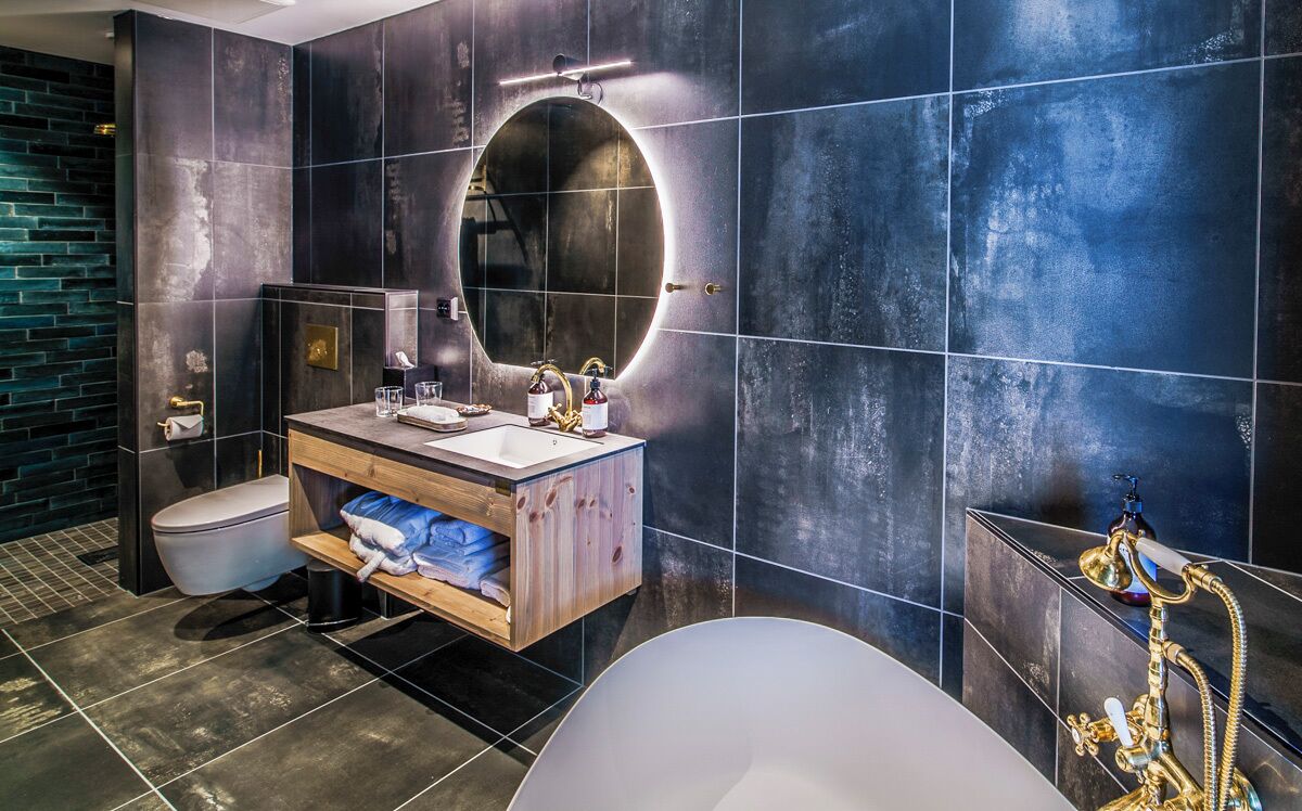 A modern bathroom with a round mirror, dark tiles, a wooden vanity, a toilet, and a bathtub with a gold faucet.