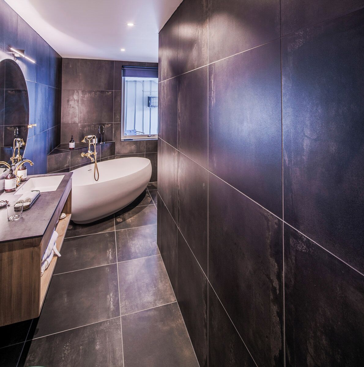 A modern bathroom with a freestanding bathtub, dark tiled walls, and a vanity with a mirror and lighting.