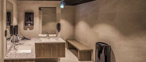Une salle de bain moderne avec un grand miroir, deux lavabos, un banc et une poubelle.