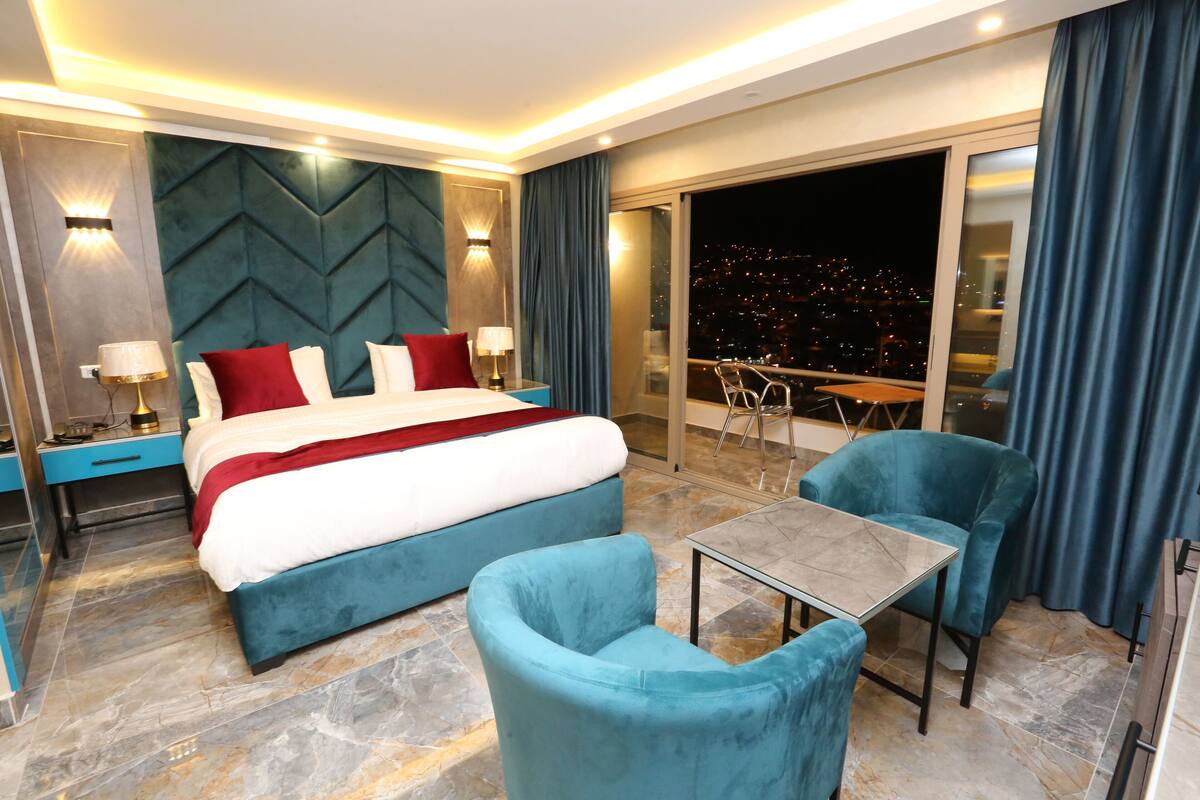 A modern hotel room with a large bed, two blue armchairs, a small table, and a balcony with a city view at night.