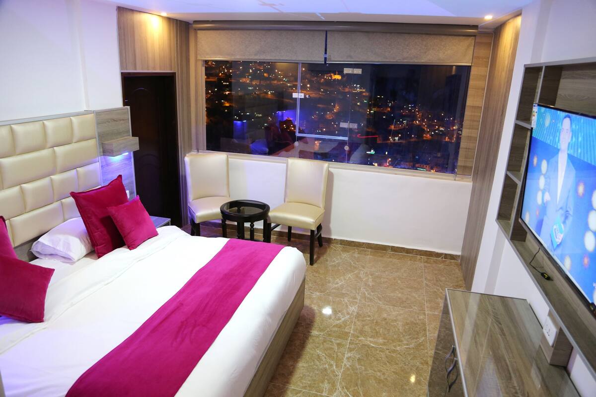 A hotel room with a large bed, a TV, a city view, and a small seating area.