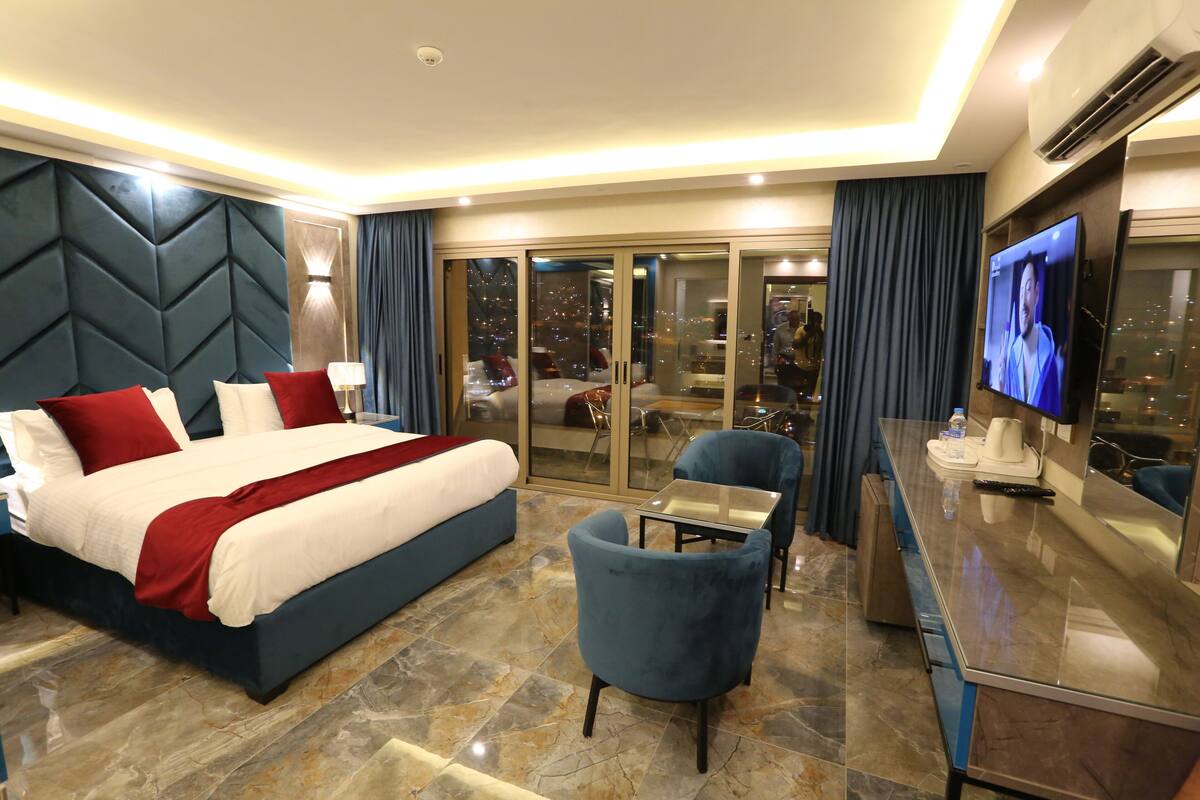 A hotel room with a large bed, a desk with a TV, and a view of the city at night.