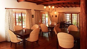 A dining area with wooden tables and chairs, large windows with curtains, and a wooden ceiling. - Mongoose Manor (Gqeberha (Port Elizabeth))