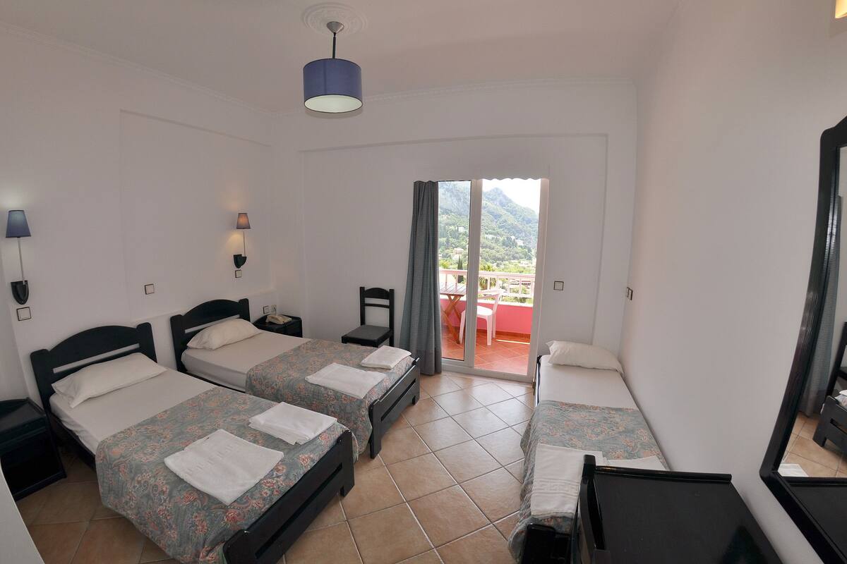 A hotel room with two beds, a desk, a chair, and a balcony with a view of the mountains.
