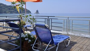 A deck with two blue lounge chairs and an orange umbrella, overlooking a calm sea and rocky coastline. - The Pink Palace (Corfu)