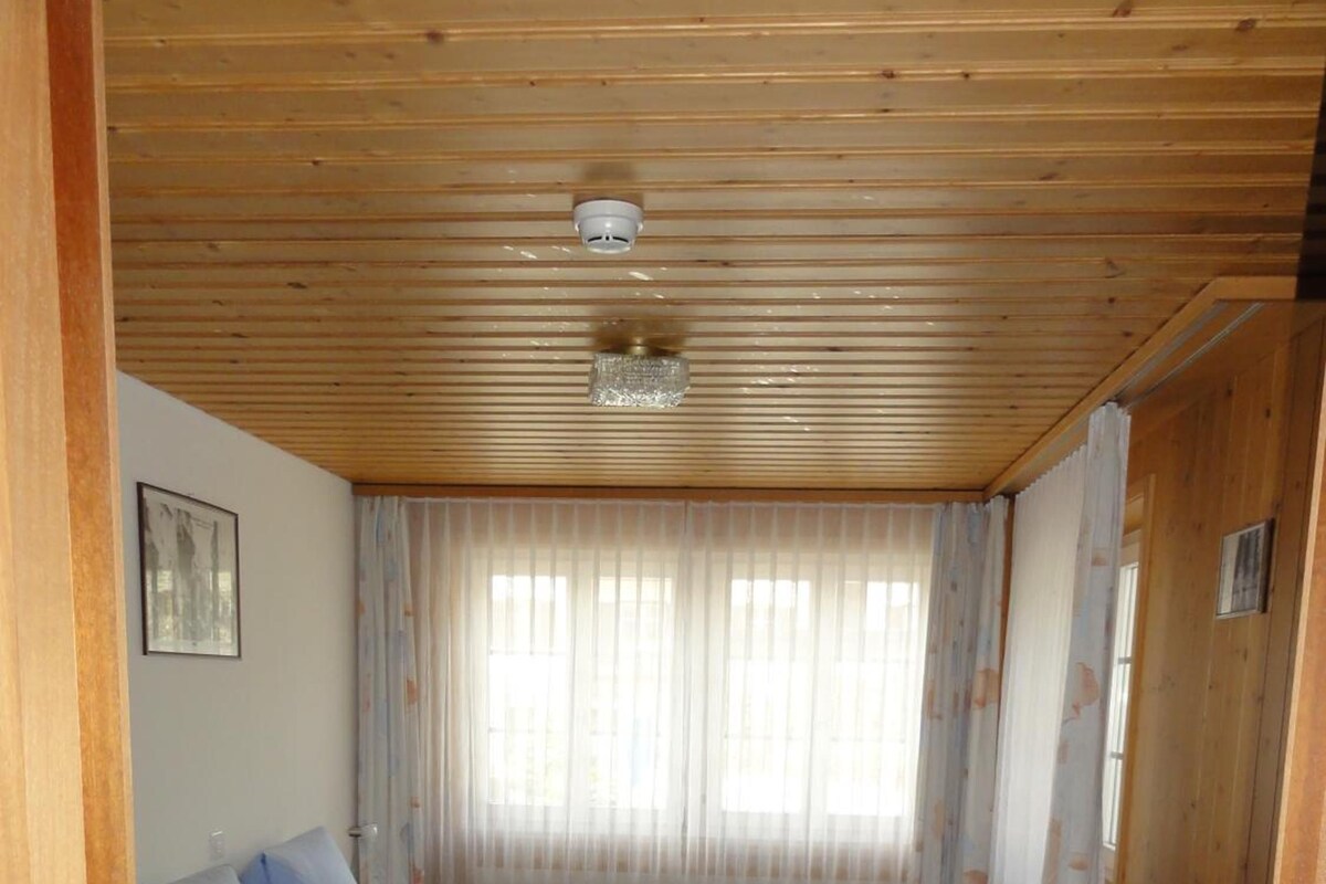 A room with a wooden ceiling, a bed with blue bedding, a chair, and a window with curtains.