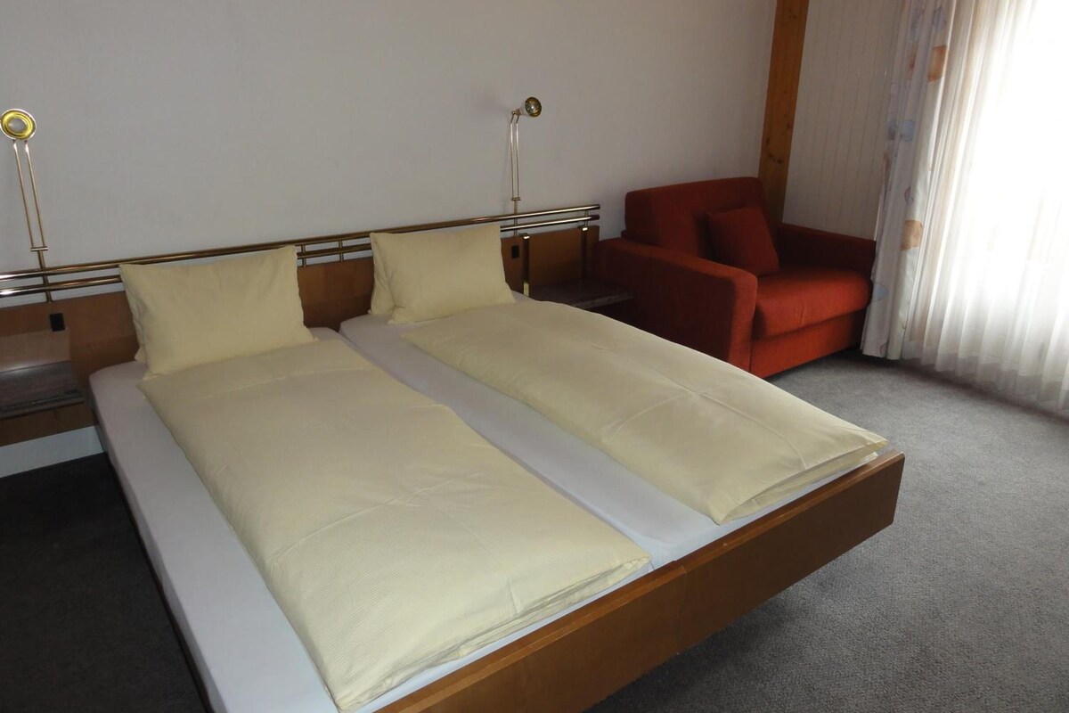 A double bed with a wooden frame and two pillows, a red chair, and a bedside table with a lamp.