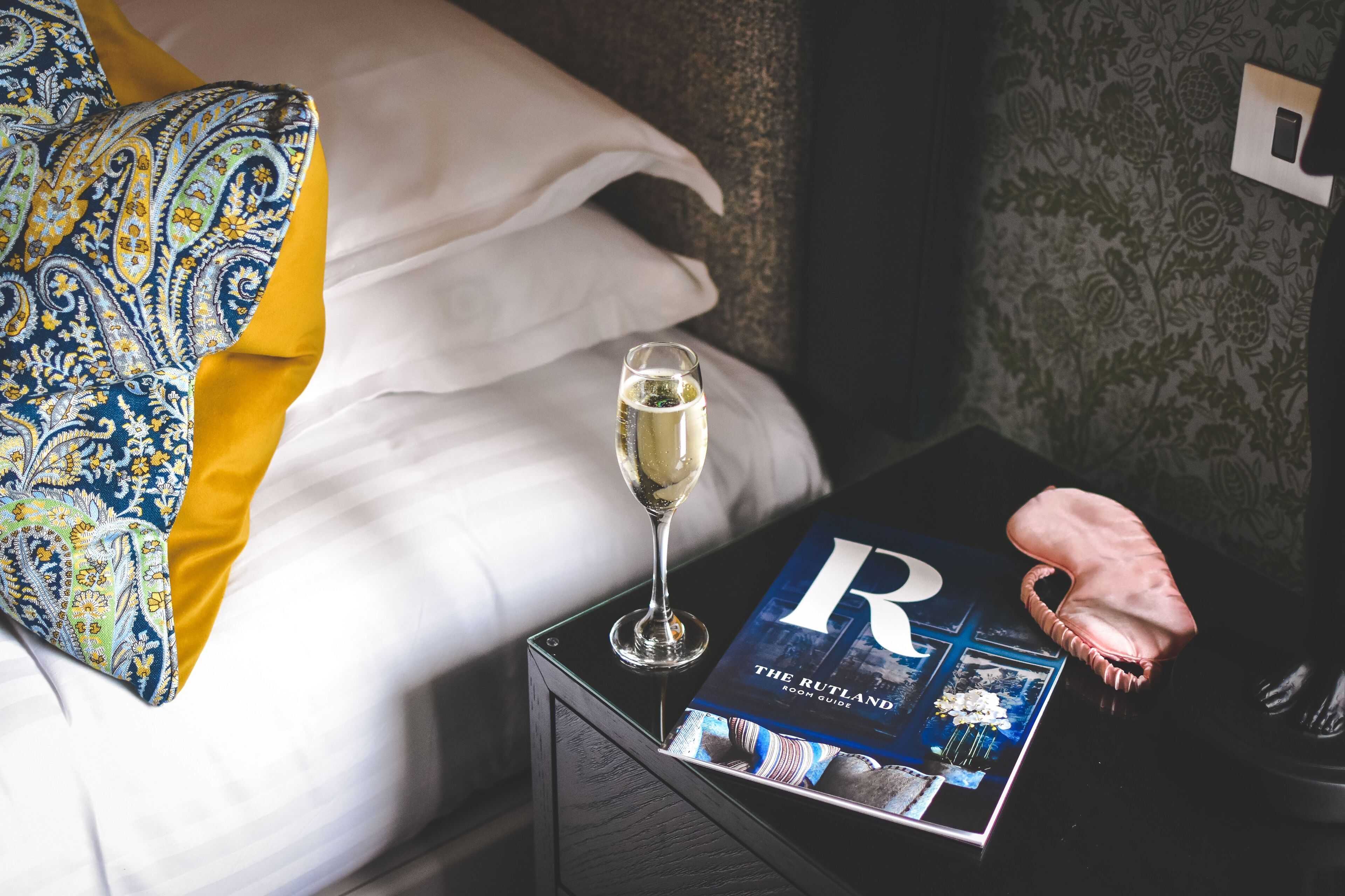 A neatly made bed with a patterned pillow, a glass of champagne, a magazine titled "The Rutland Room Guide," and a small oven mitt on a bedside table.