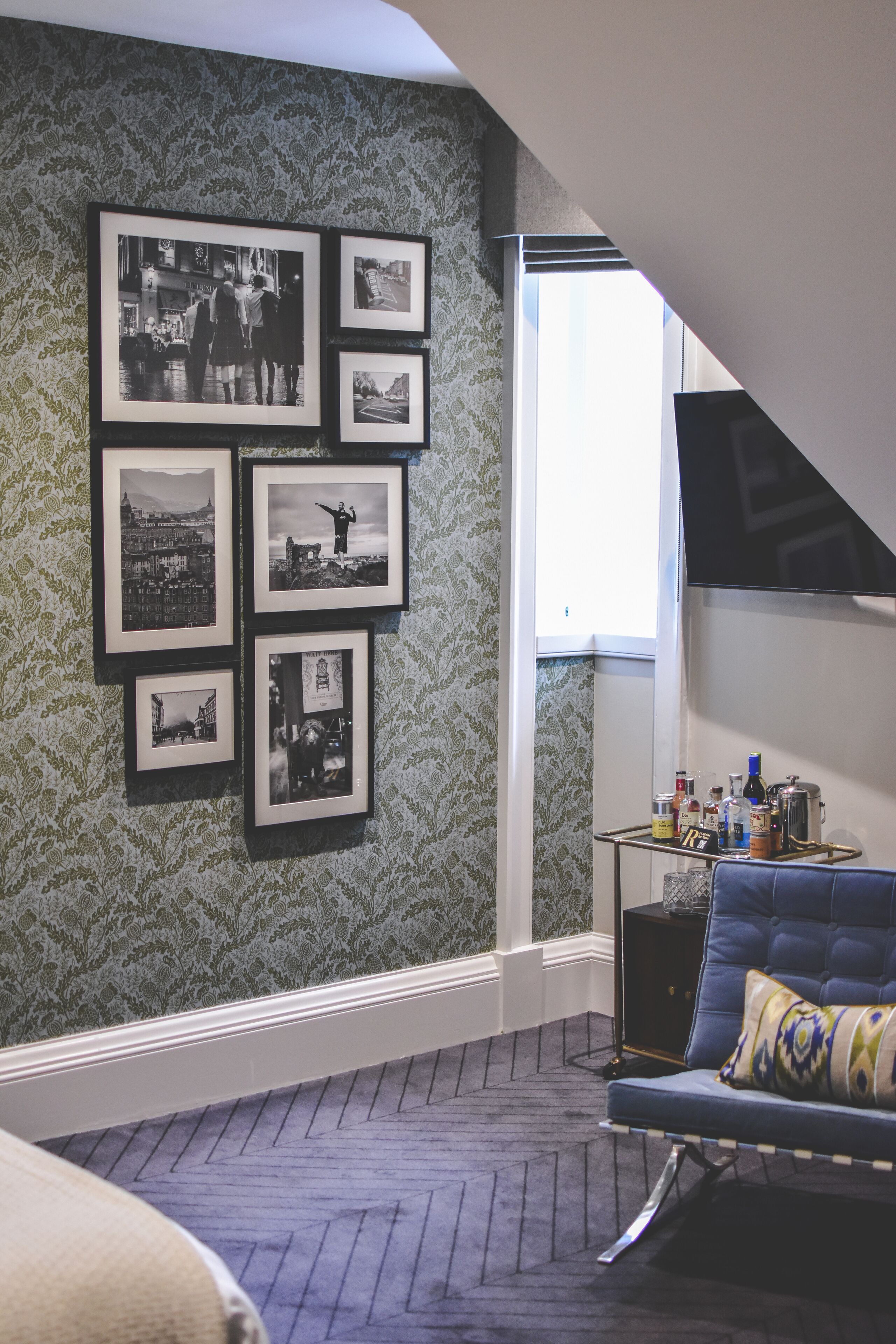 Um quarto com piso em padrão herringbone, um sofá azul com almofada estampada, e uma parede decorada com fotografias em preto e branco emolduradas.