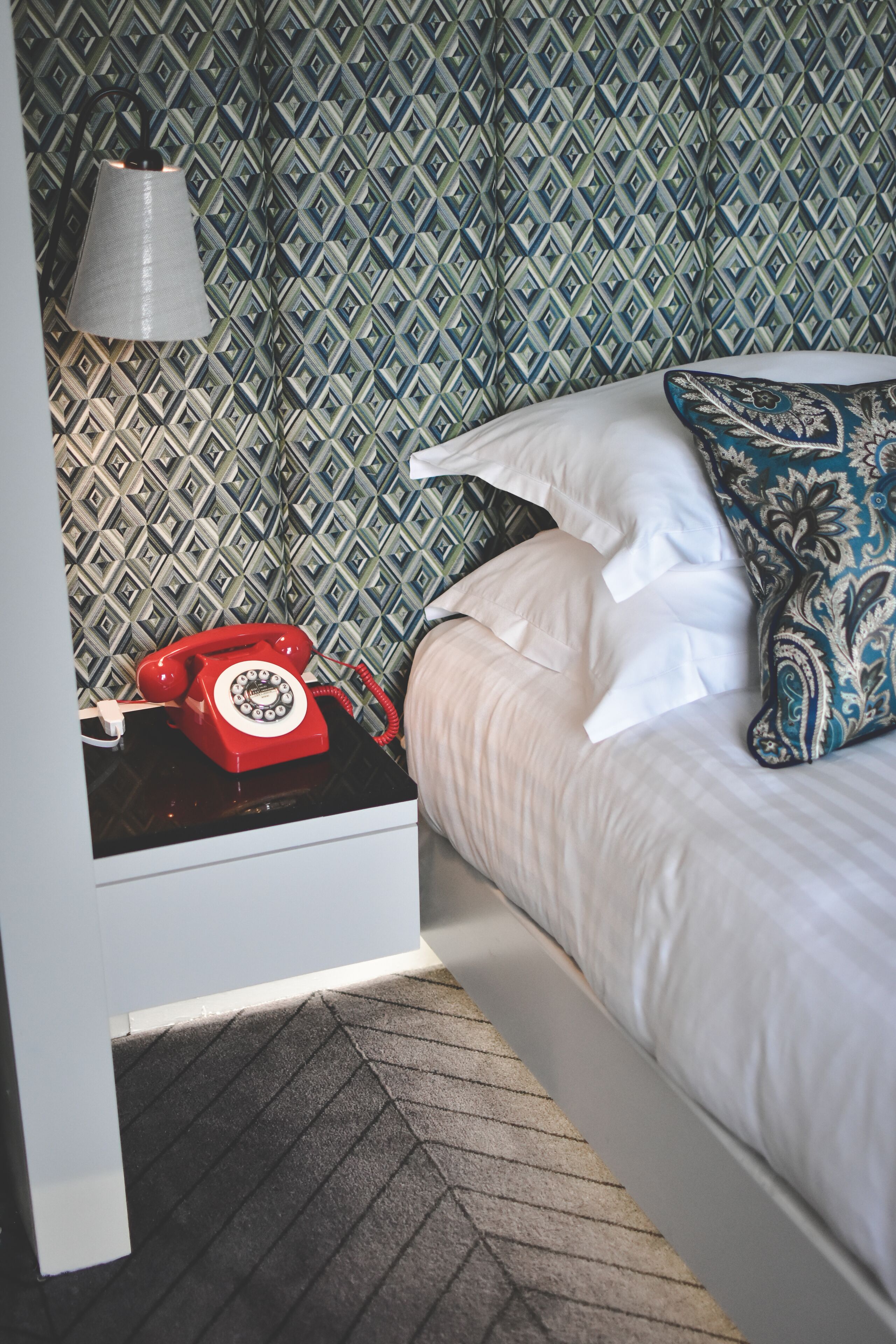 A hotel room with a patterned wallpaper, a red rotary phone on a bedside table, a white bed with a blue patterned pillow, and a wall-mounted lamp.