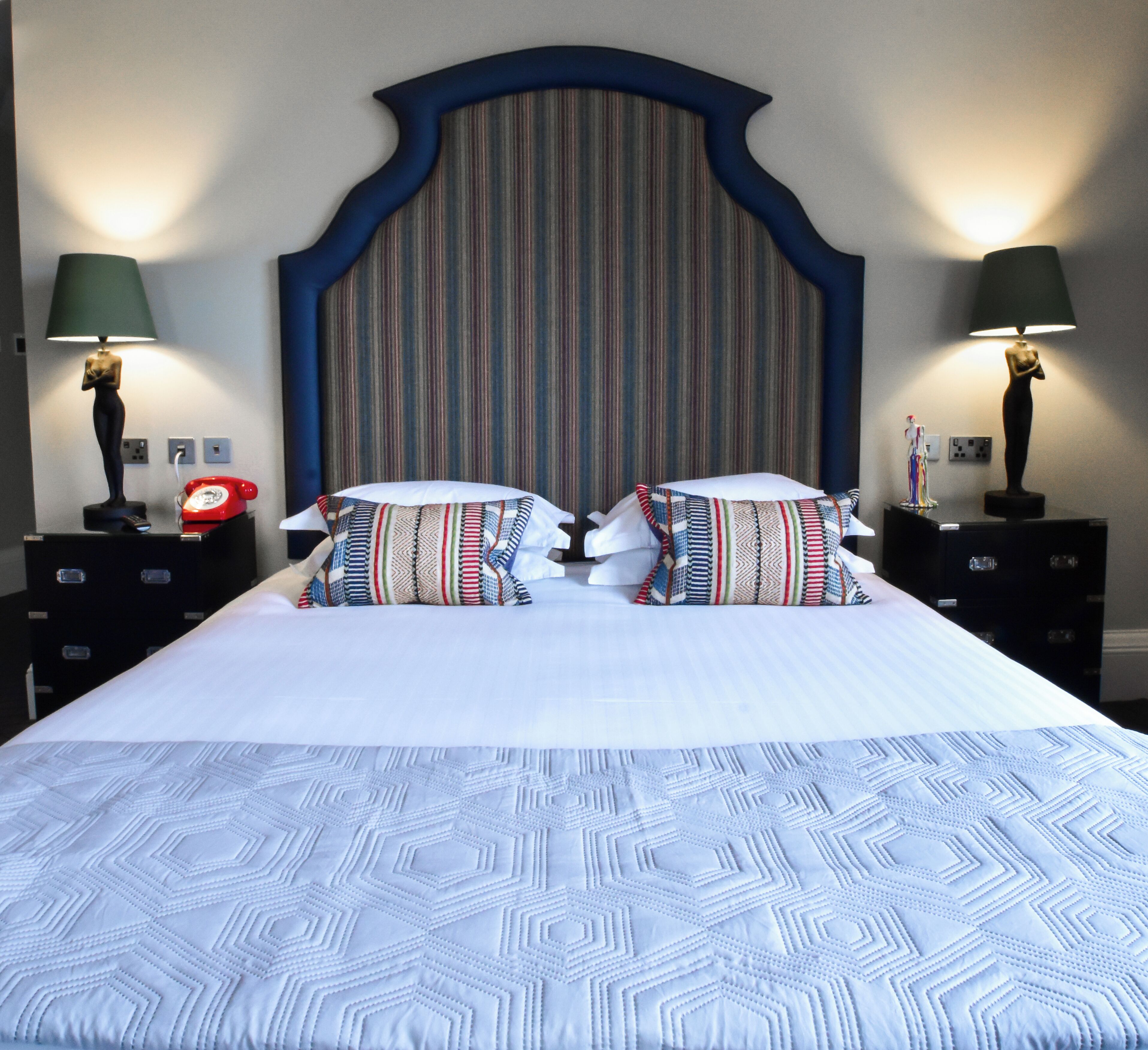 Une chambre d’hôtel avec un grand lit, une tête de lit à rayures, deux tables de chevet avec des lampes et un téléphone.