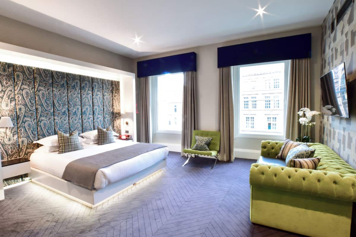 A modern hotel room with a large bed, a green armchair, a sofa, and a TV mounted on the wall.