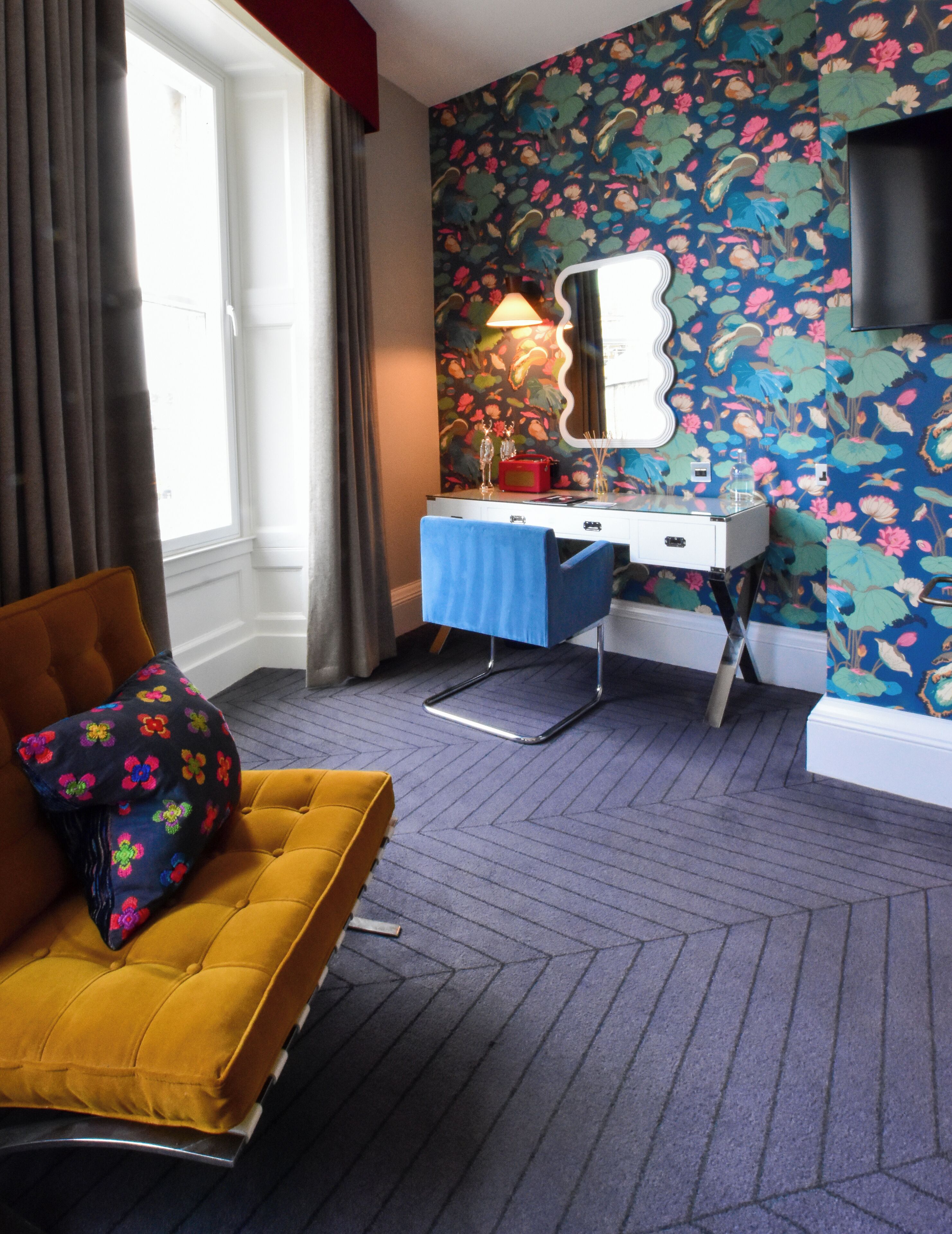 A room with a floral wallpaper, a desk with a mirror, a blue chair, and a yellow sofa.