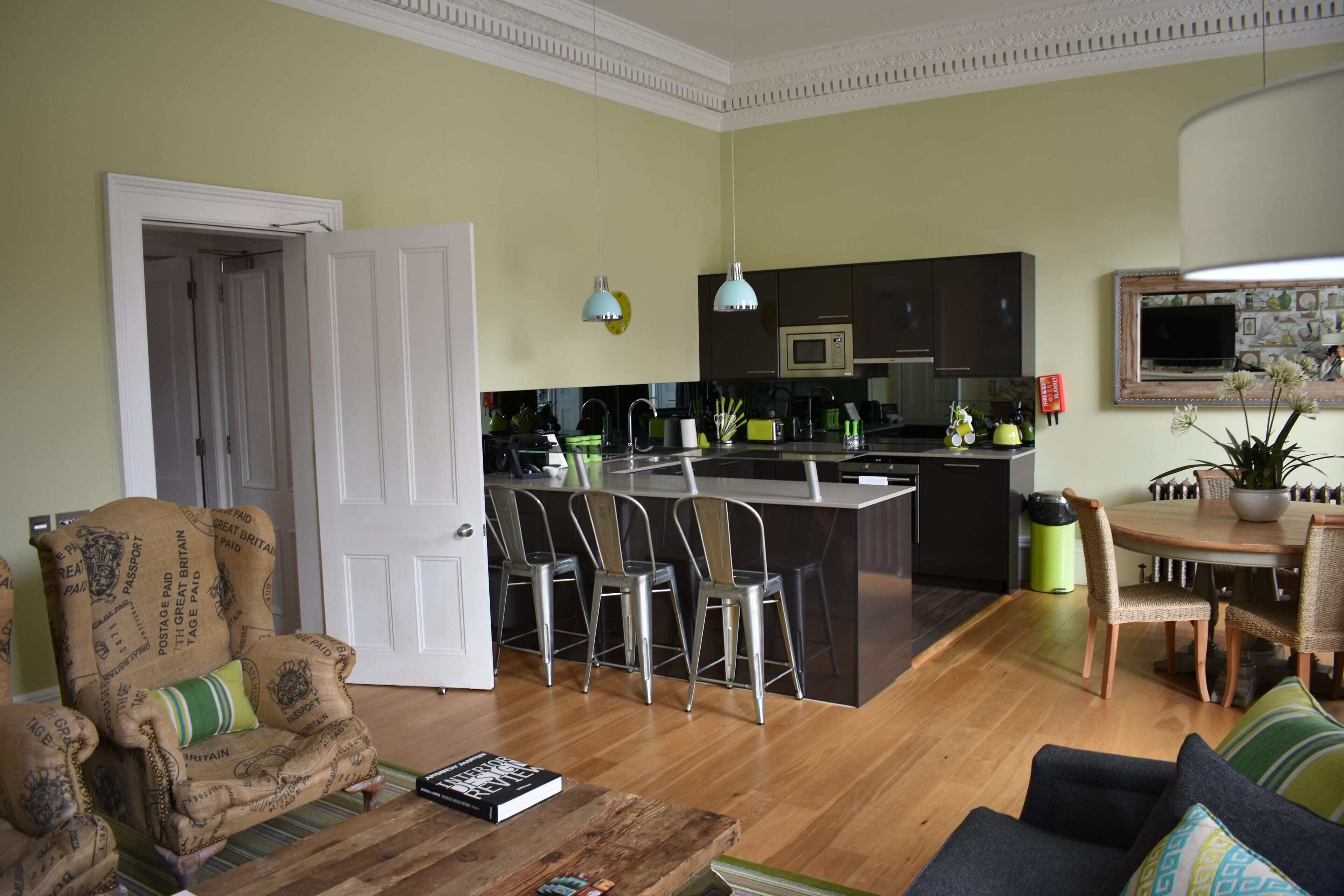 A living room with a sofa, a coffee table, and a patterned armchair. There is a kitchen area with a dining table and chairs.