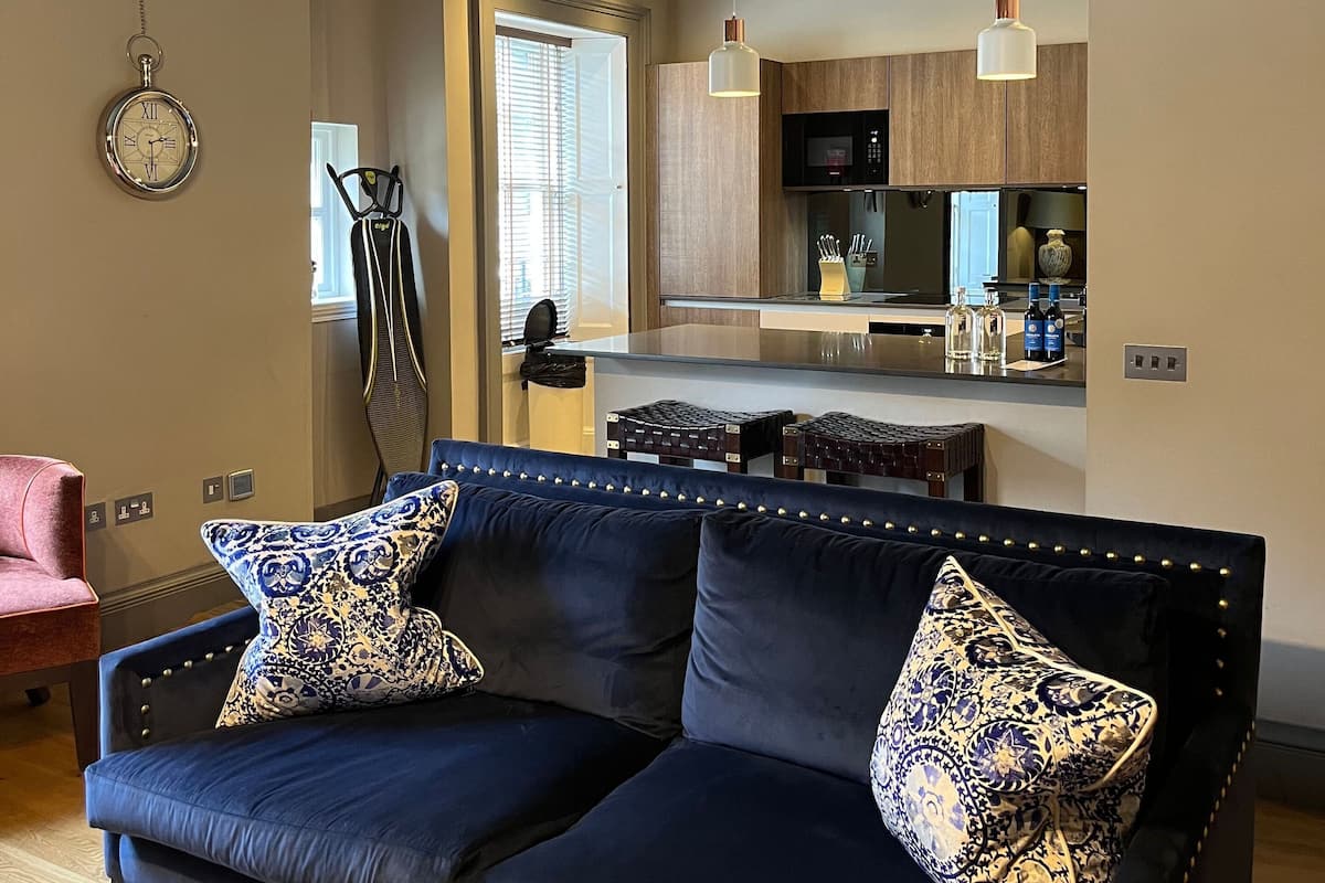 A modern living room with a dark blue sofa, patterned cushions, and a wooden floor. A kitchen area is visible in the background with wooden cabinets and a microwave.