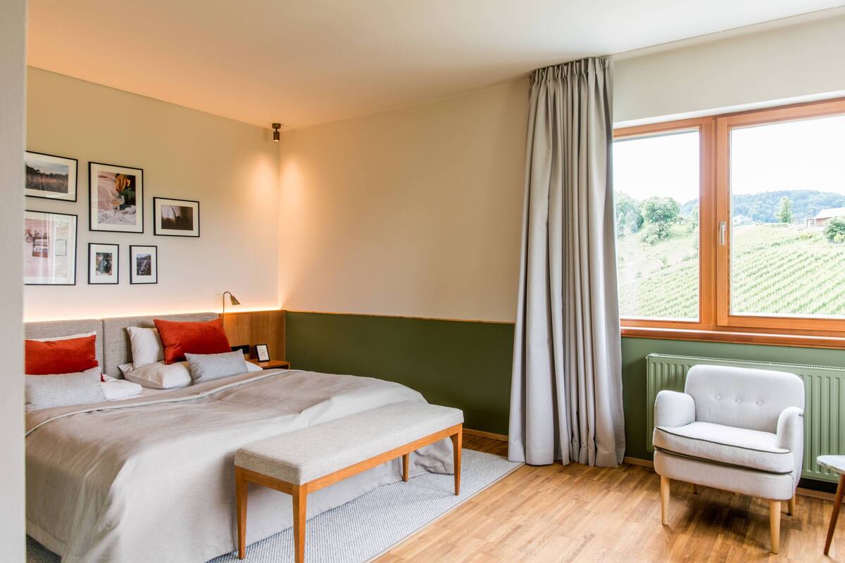 A bedroom with a bed, a chair, a window with curtains, and framed pictures on the wall.