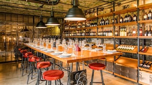 Una larga mesa dispuesta para cenar, con sillas rojas, botellas de vino en estantes y lámparas colgantes.
