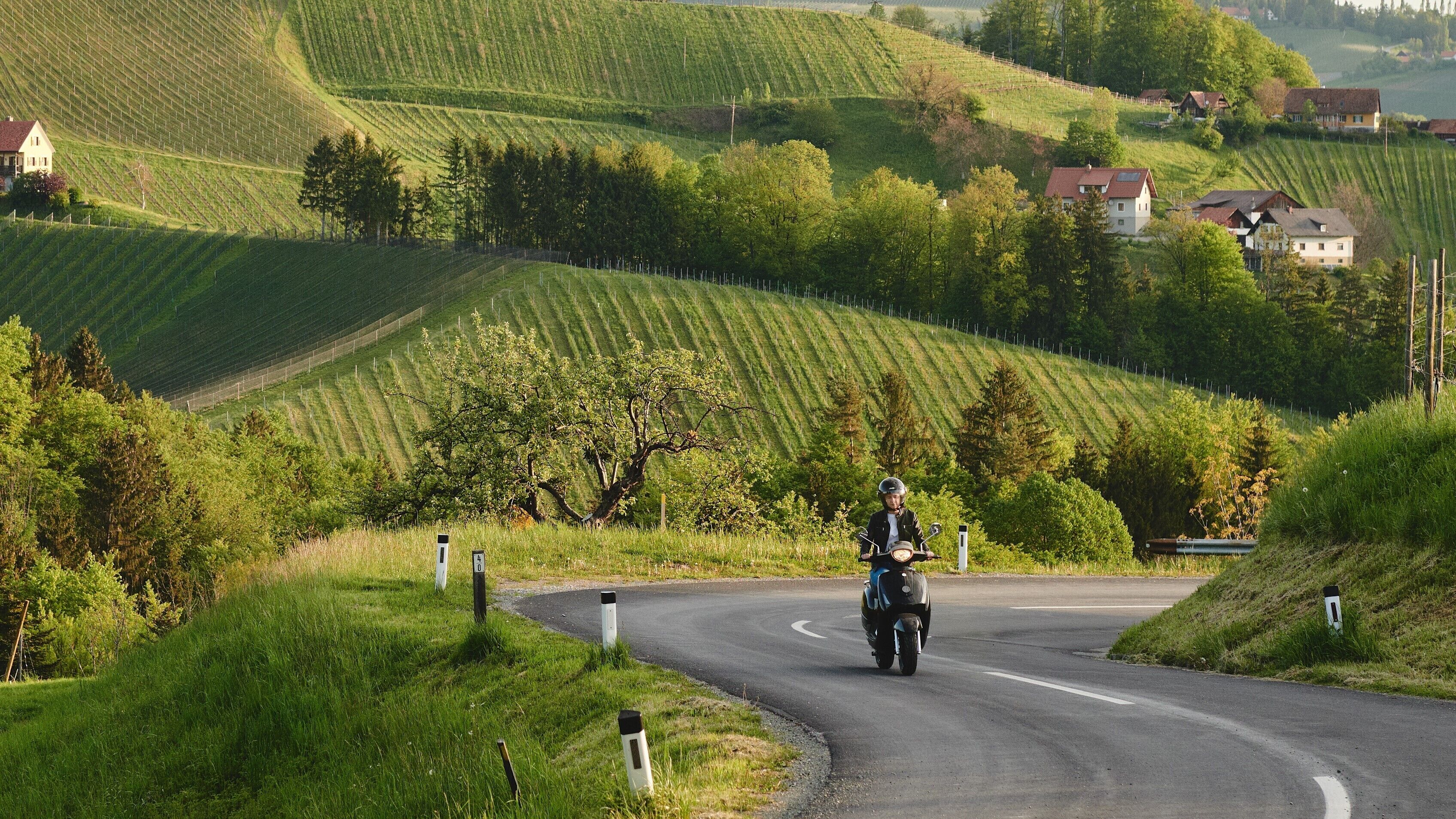 A motorcyclist on a winding road surrounded by lush green hills and scattered houses.