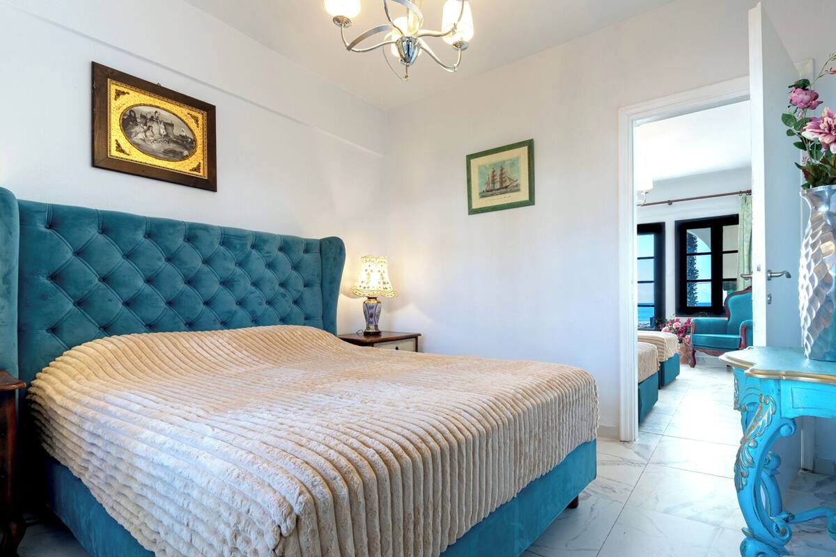 A bedroom with a large blue upholstered headboard, a bed with a beige quilt, a nightstand with a lamp, and two framed pictures on the wall.
