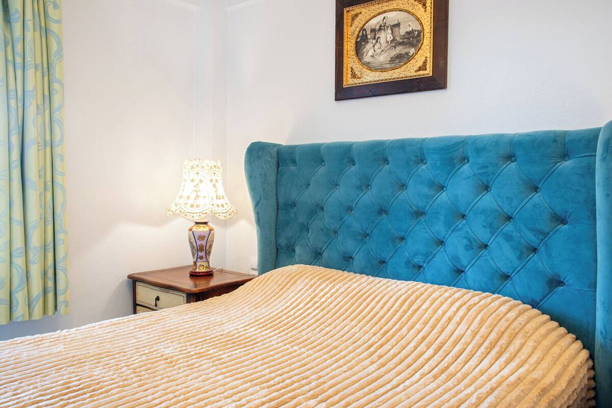 A bedroom with a tufted blue headboard, a patterned bedspread, a bedside table with a lamp, and a framed picture on the wall.