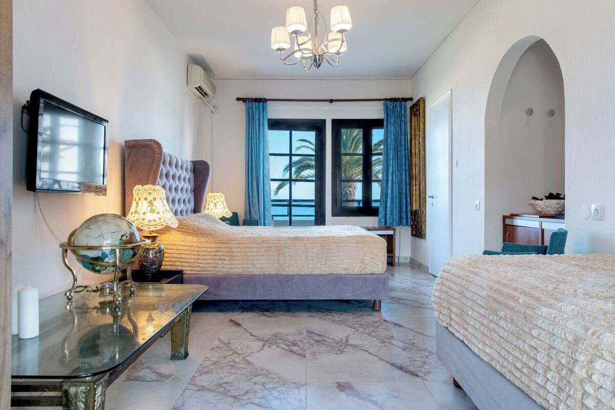 A hotel room with a bed, a desk with a globe and lamp, a TV, and a view of palm trees through the window.