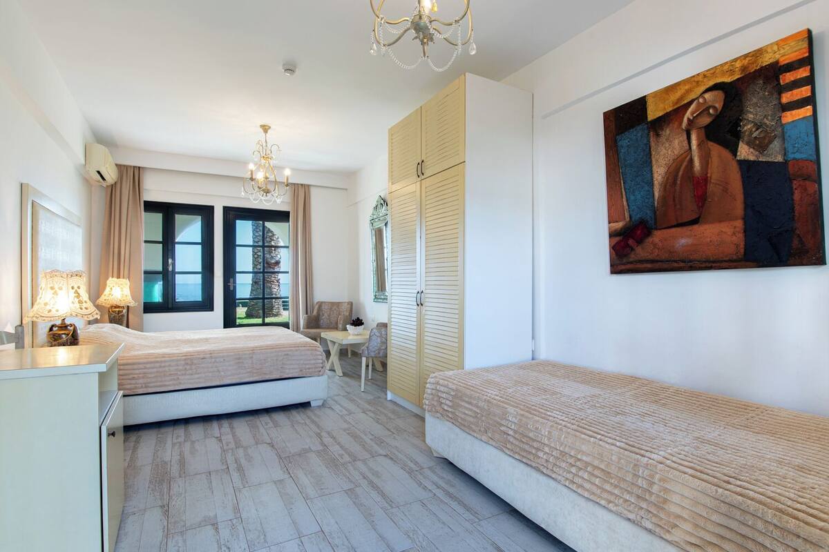 A hotel room with two beds, a chandelier, a painting, and a view of the sea through the window.