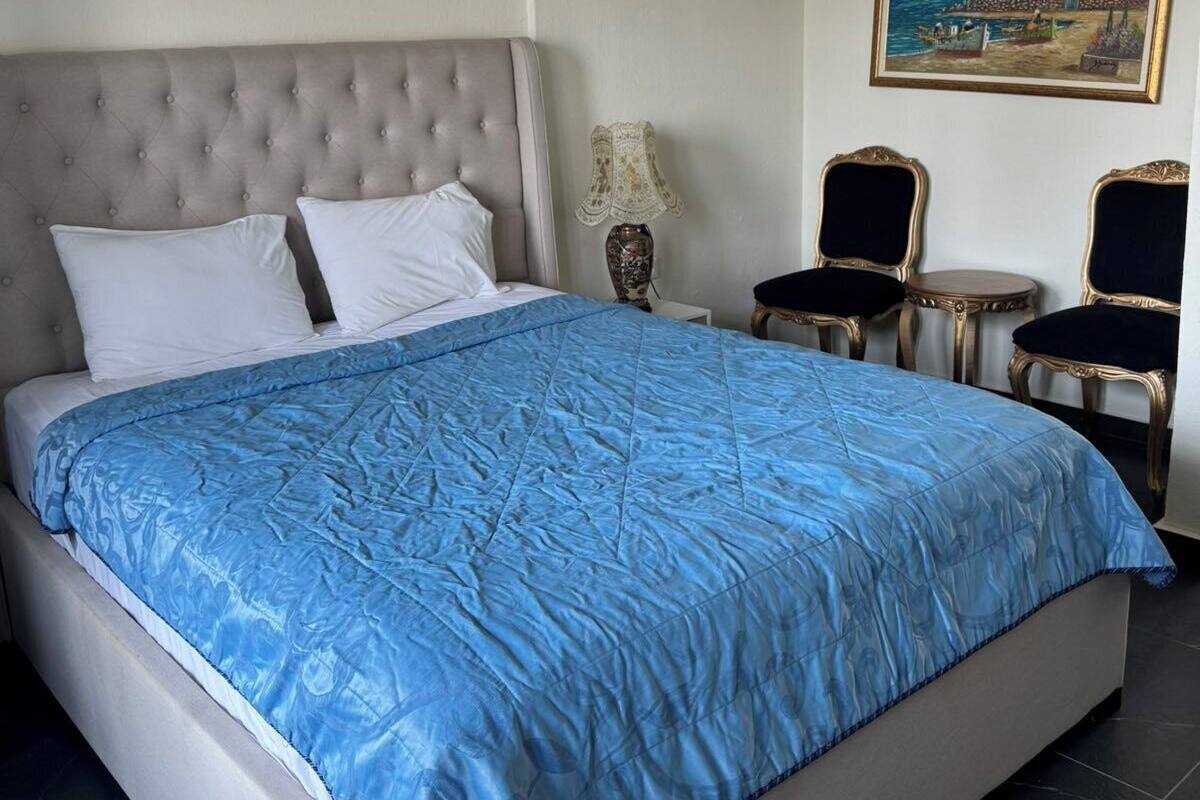 A neatly made bed with a blue quilt, a grey upholstered headboard, and two chairs with ornate backs.