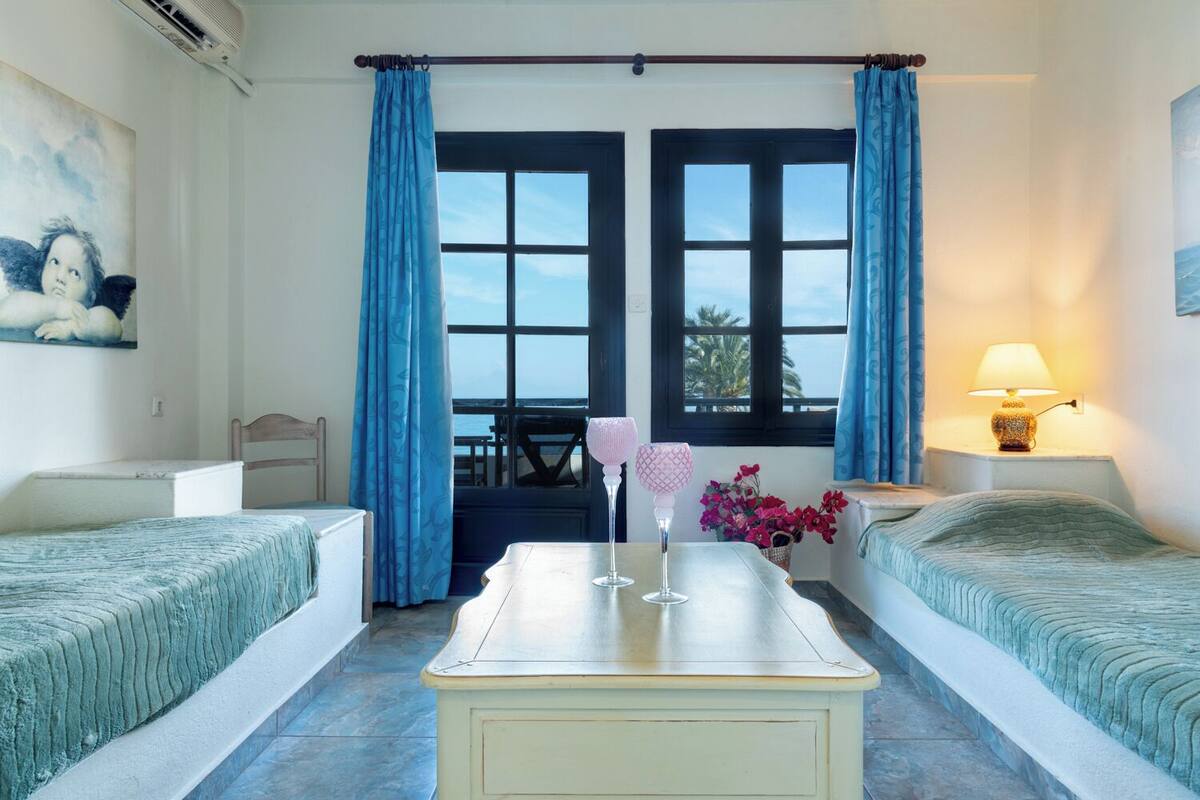 A room with two beds, a table, a lamp, and a view of the sea through large windows.