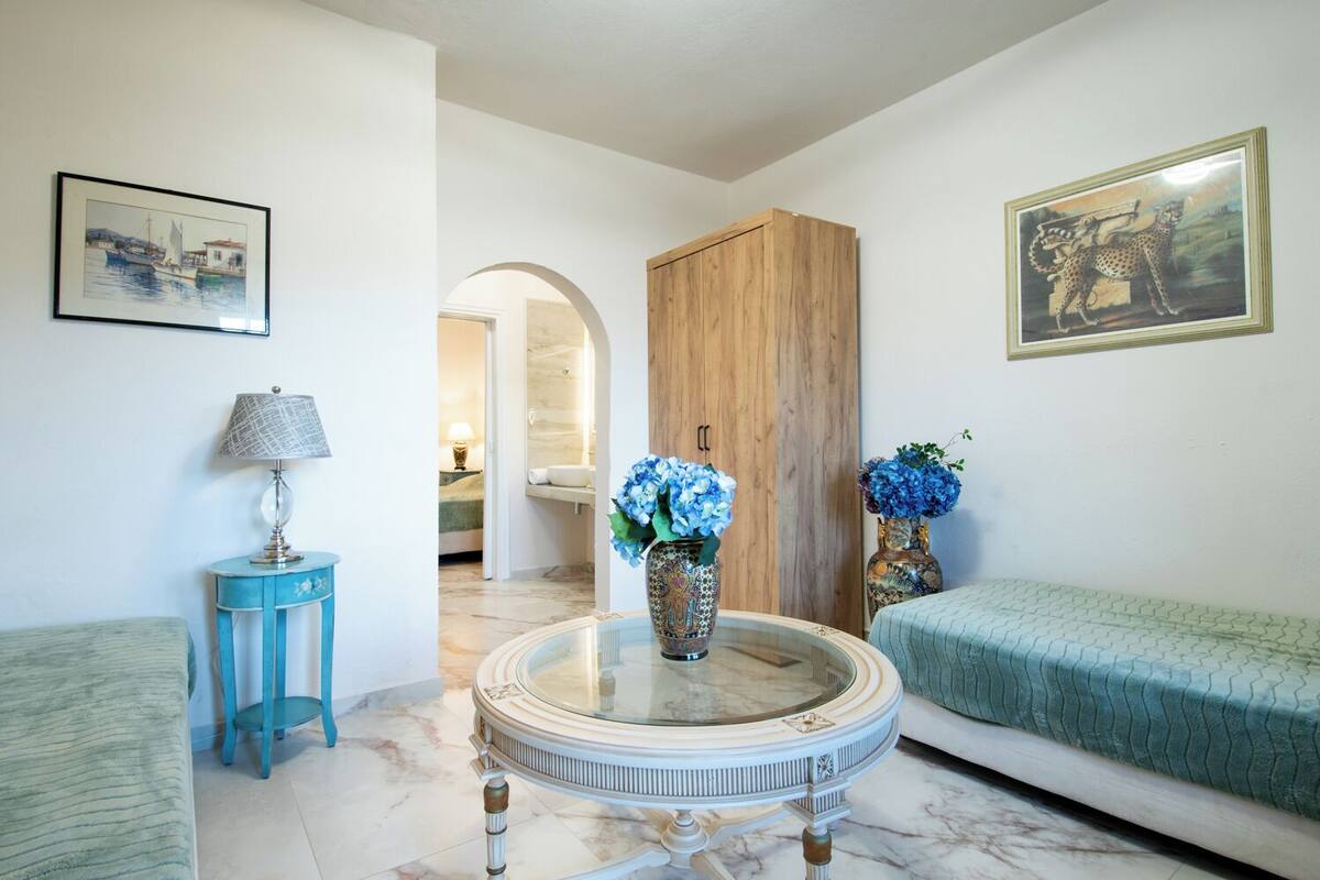 A room with a round marble table, two vases with blue flowers, a blue side table, a green sofa, and a wooden wardrobe.