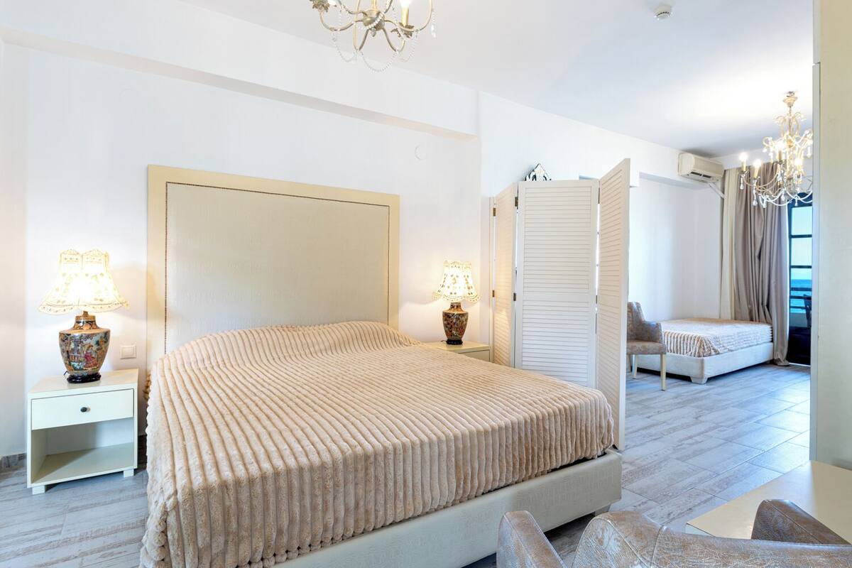 A modern bedroom with a large bed, bedside lamps, a wardrobe, and a view of the sea.