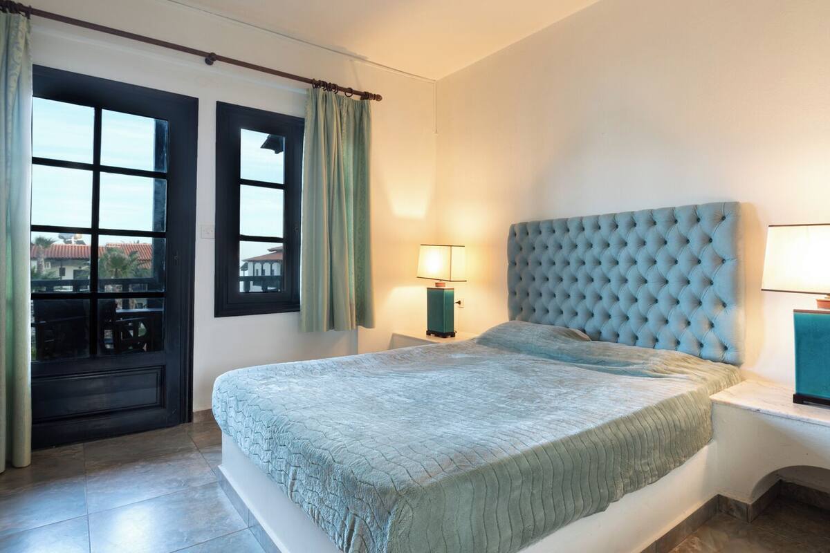 A bedroom with a large bed, a window with curtains, a nightstand with a lamp, and a view of buildings outside.