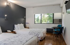 A hotel room with two beds, a desk with a chair, a window with a view of buildings, and a wall-mounted TV.