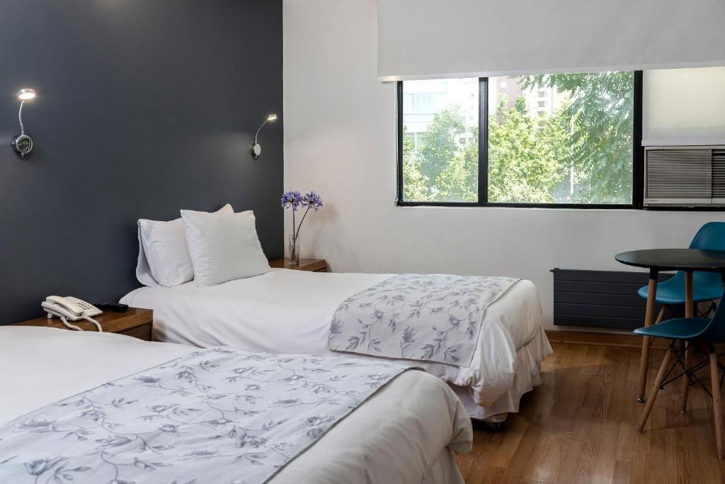 Habitación de hotel con dos camas, una mesita, sillas, un teléfono en la mesita de noche y una ventana con vistas a árboles.
