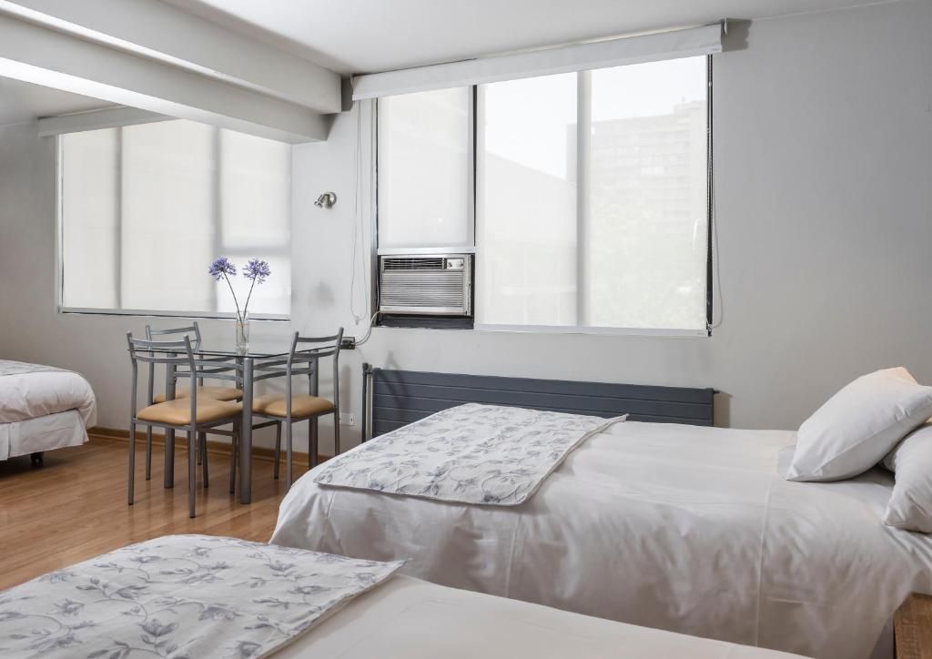 A hotel room with two beds, a dining table with chairs, a radiator, and a window with blinds.