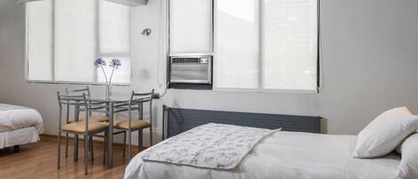 A hotel room with two beds, a dining table with chairs, a radiator, and a window with blinds.