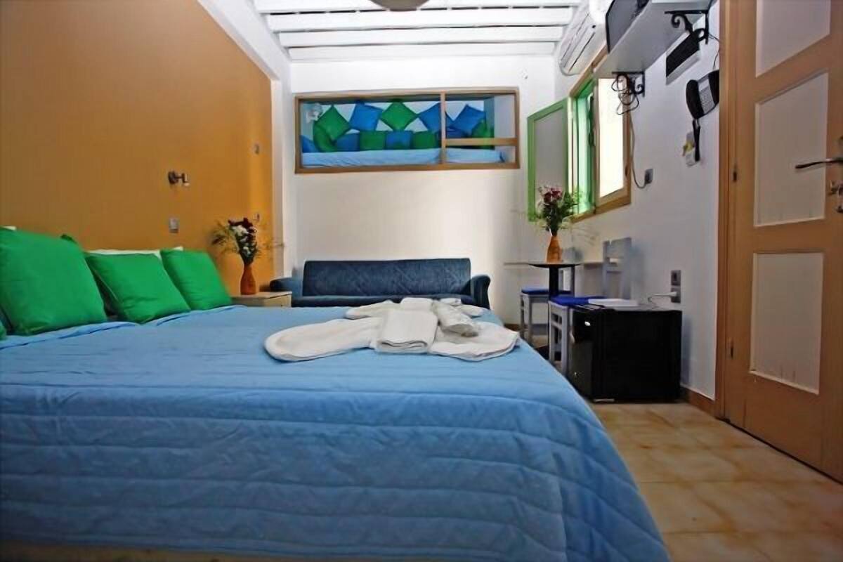 A hotel room with a bed, a desk, a chair, a small table, a mirror, and a window with a geometric painting.