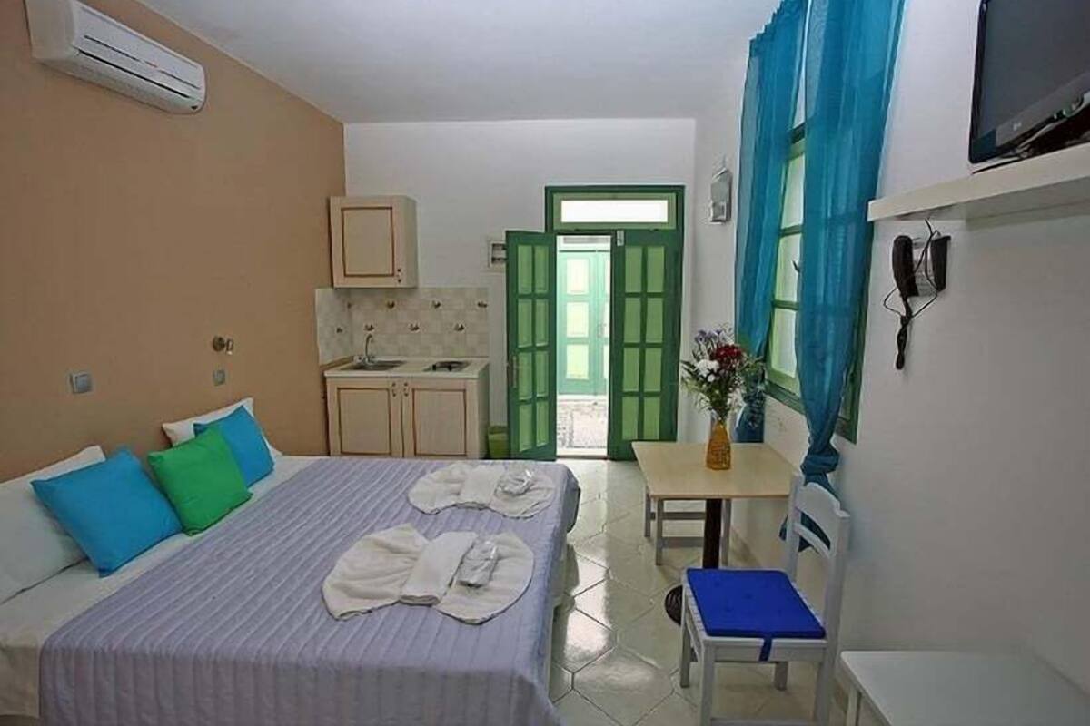 A compact hotel room with a bed, a small table, a chair, a kitchenette, and a bathroom visible through an open door.