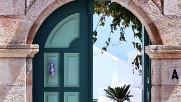 A stone archway leading to a room with a green door, a potted plant, and a bed with a white headboard. - Xenones Lindos (Rhodes)