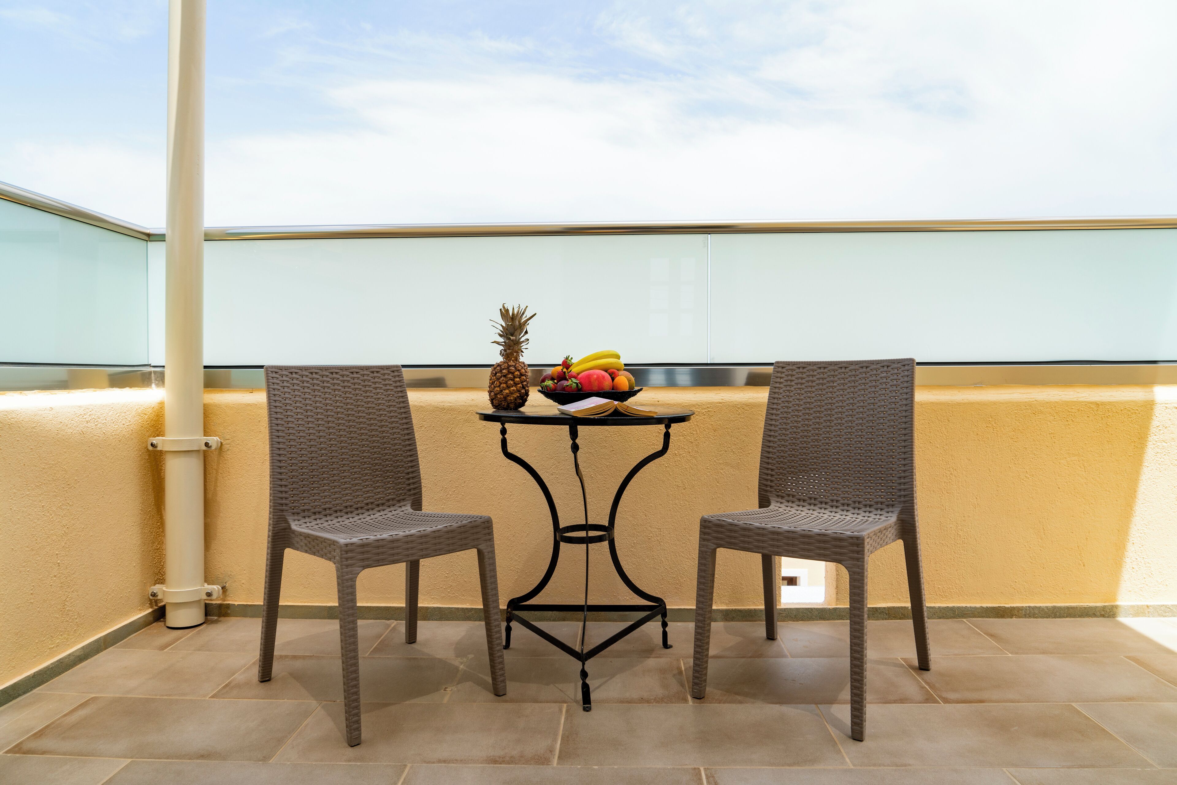 A balcony with two wicker chairs, a small table with a fruit bowl, and a clear view of the sky.