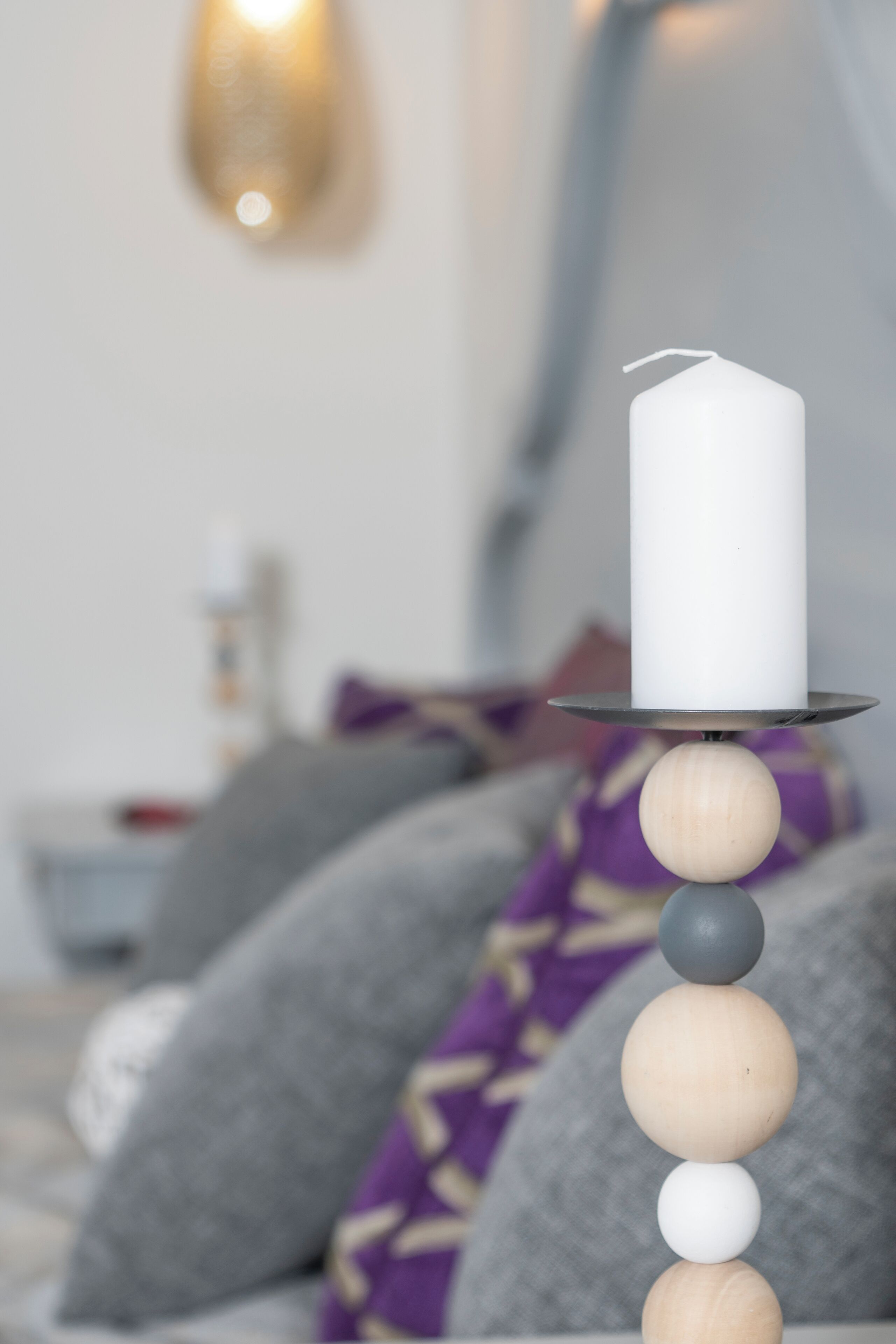 A white candle on a wooden stand with a metal base, set against a blurred background of a bed with pillows and a wall-mounted light fixture.