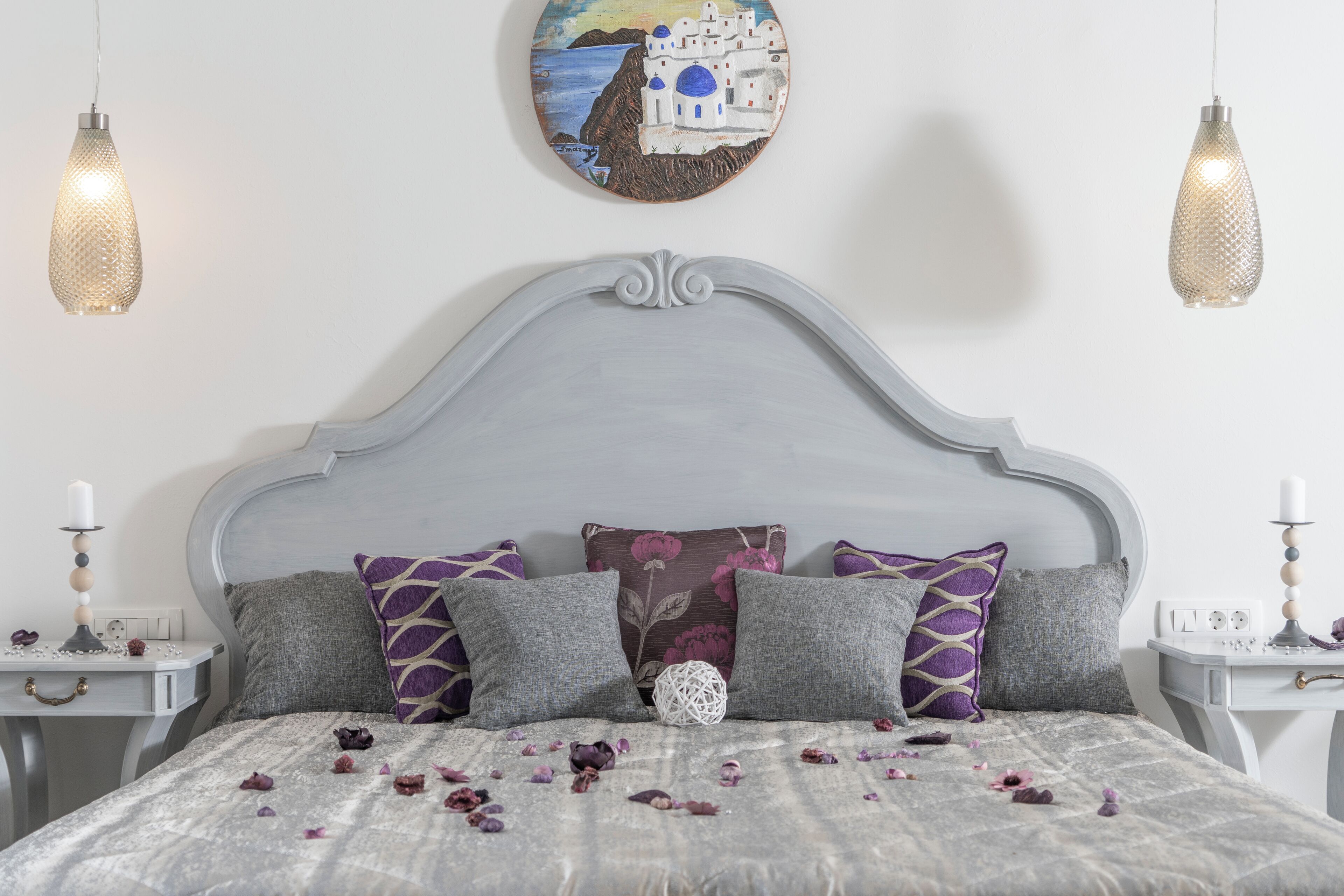 A bedroom with a grey upholstered headboard, a bed with a grey quilt and purple and grey pillows, a white wall with a circular painting, and two bedside tables with candles and a decorative item.