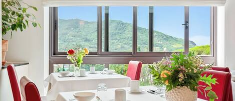 A dining area with tables set for two, white tablecloths, and a view of green hills through large windows.