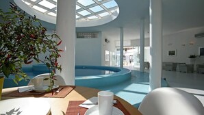 Un espace piscine intérieur moderne doté d’un toit en verre, d’un coin salon avec une table préparée pour deux personnes, et offrant une vue sur la piscine.