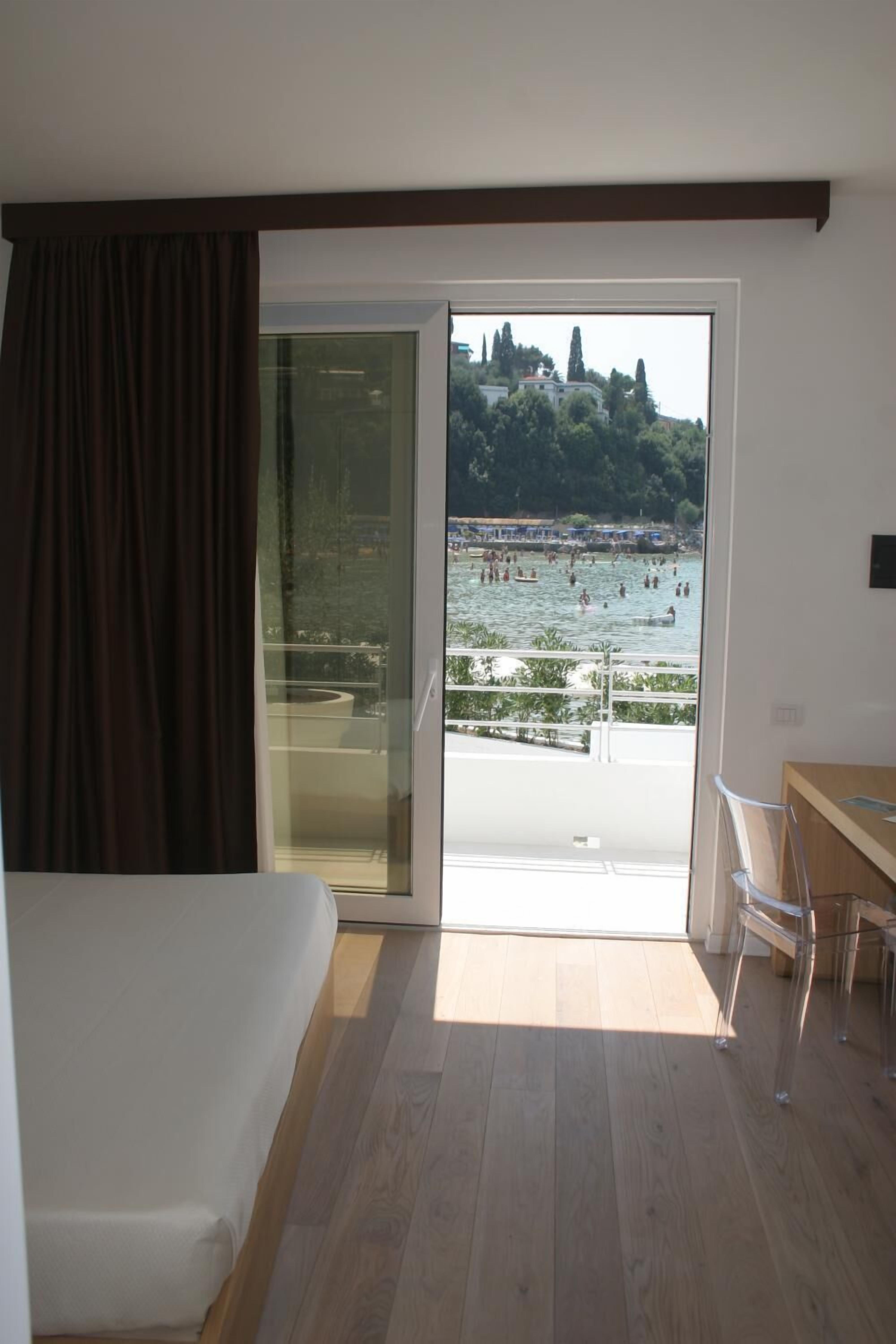 Ein Hotelzimmer mit einer großen Schiebeglastür, die auf einen Balkon mit Seeblick und badenden Personen führt.
