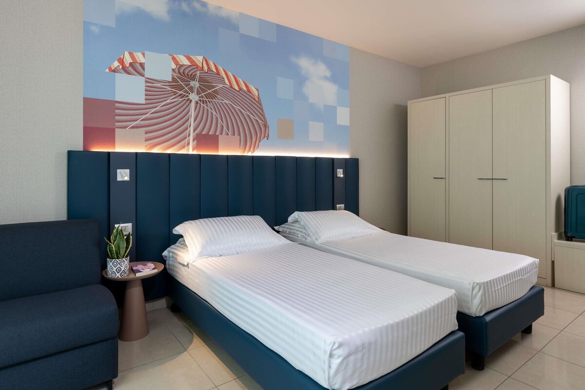 Quarto de hotel com duas camas, um sofá, um guarda-roupa e um mural na parede.