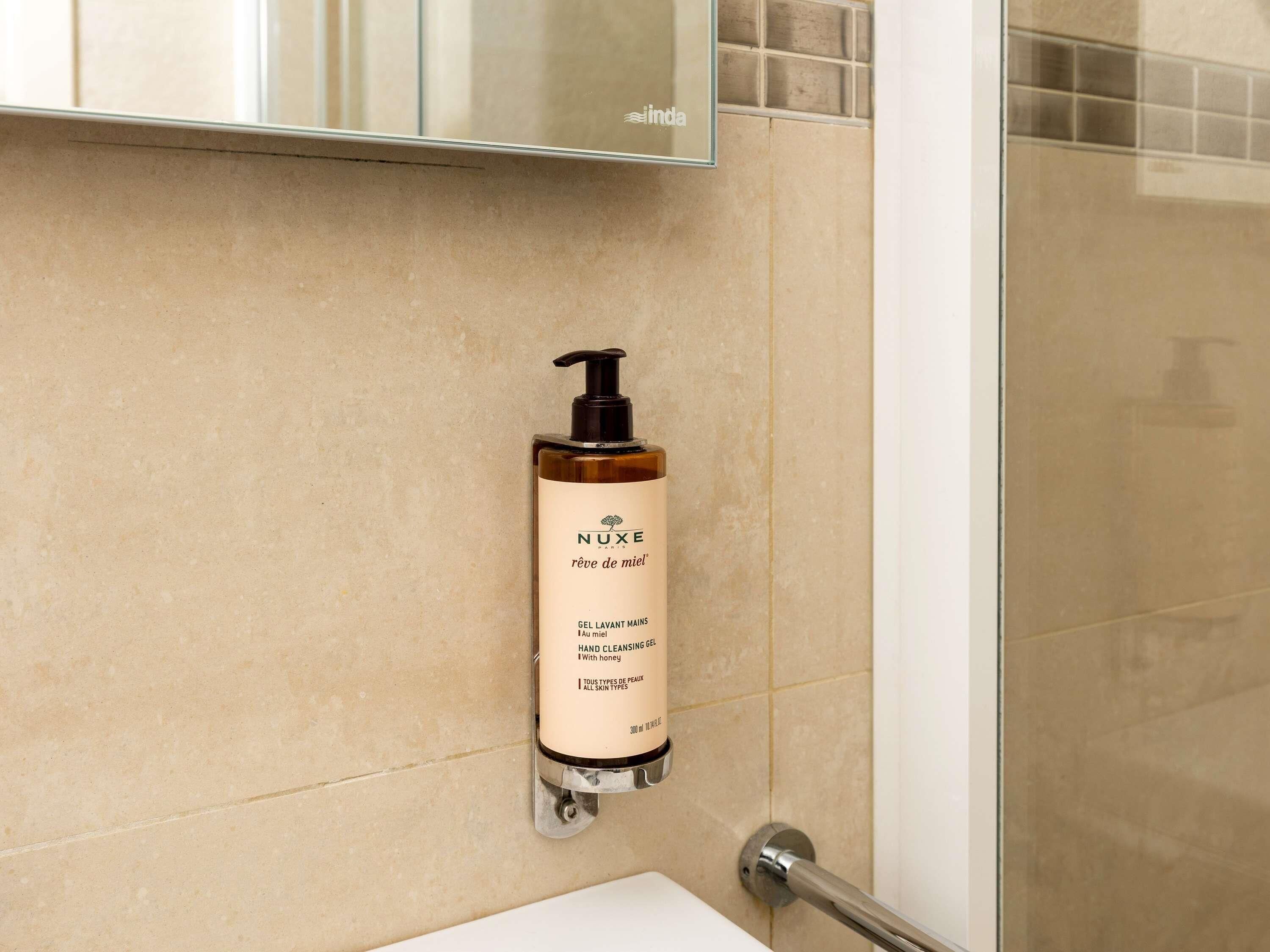 A bathroom with a hand cleansing gel dispenser mounted on the wall, a shower area, and a mirror above the dispenser.