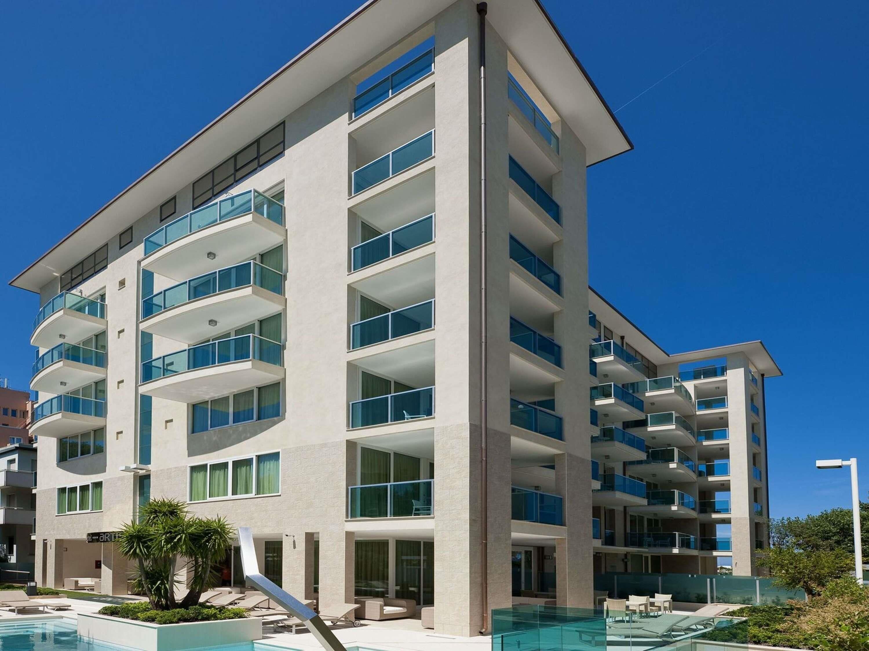 A multi-story hotel with balconies, a swimming pool, and a clear blue sky.